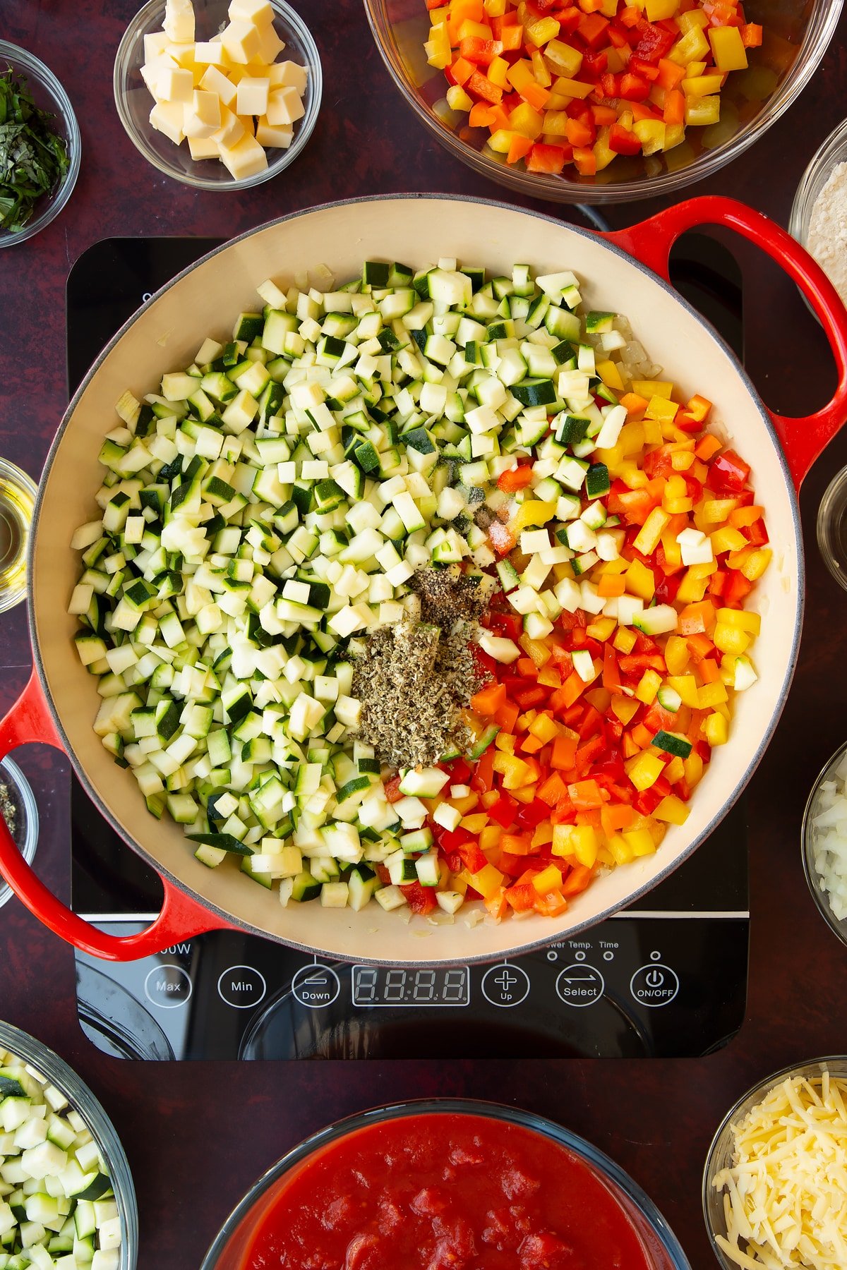 Overhead shot of finely sliced courgettes, peppers, sage, onion, salt and pepper simmering on the hob in a red pan.