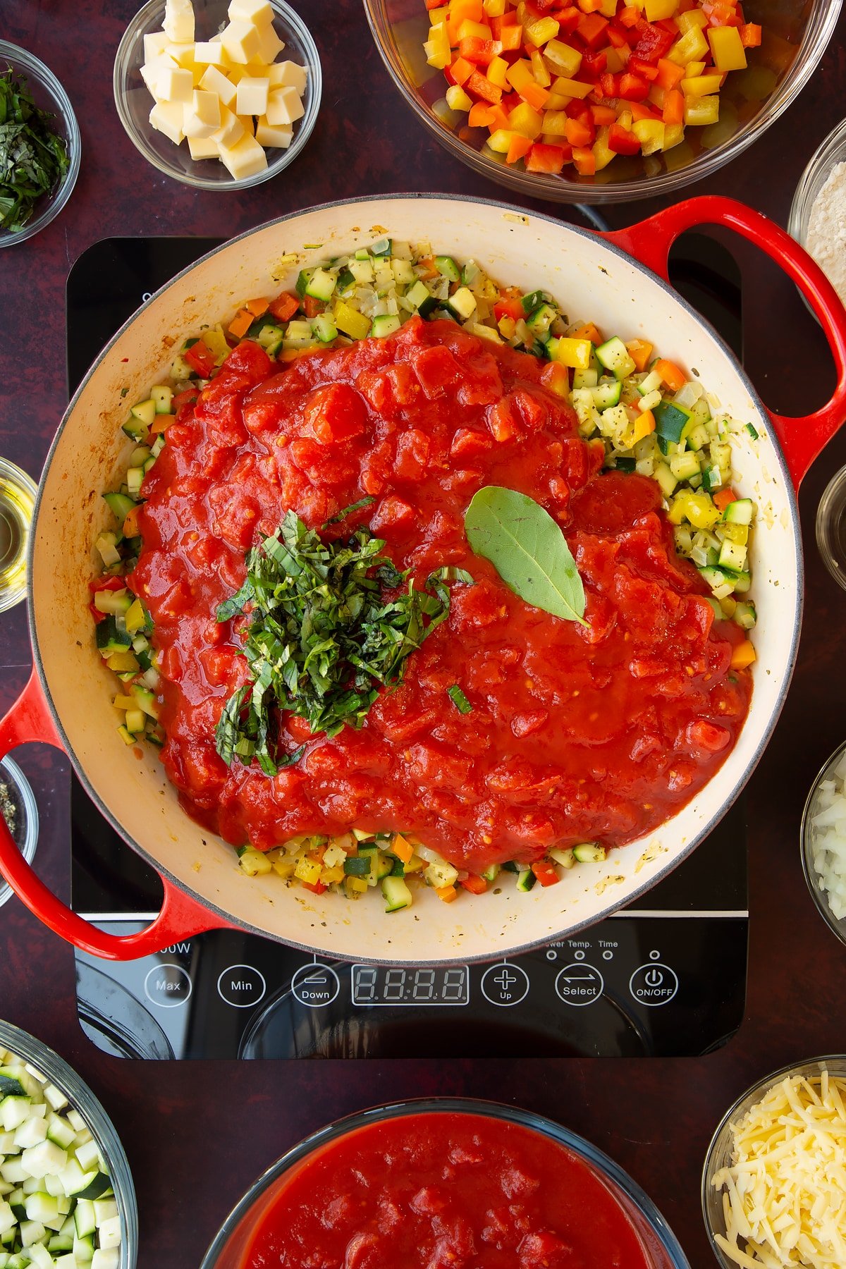 Overhead shot of finely sliced courgettes, peppers, sage, onion, salt and pepper, chopped tomatoe and basil simmering on the hob in a red pan.