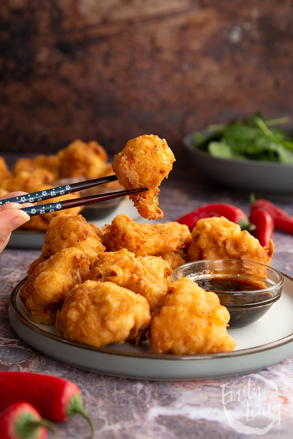 chopsticks holding a piece of buttermilk fried cauliflower over a plate full of buttermilk fried cauliflower next to BBQ dipping sauce.