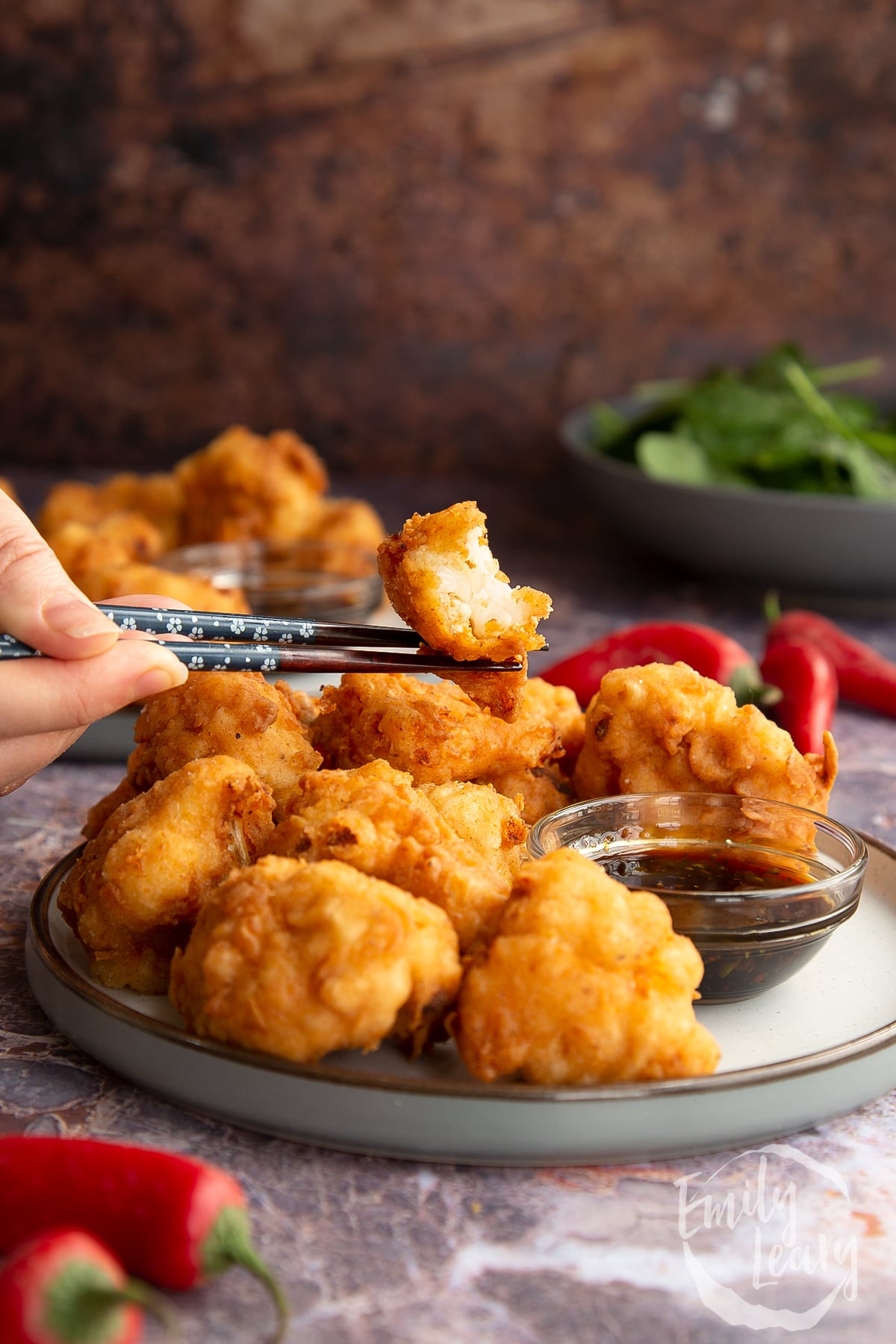 chopsticks holding a piece of buttermilk fried cauliflower over a plate full of buttermilk fried cauliflower next to BBQ dipping sauce.