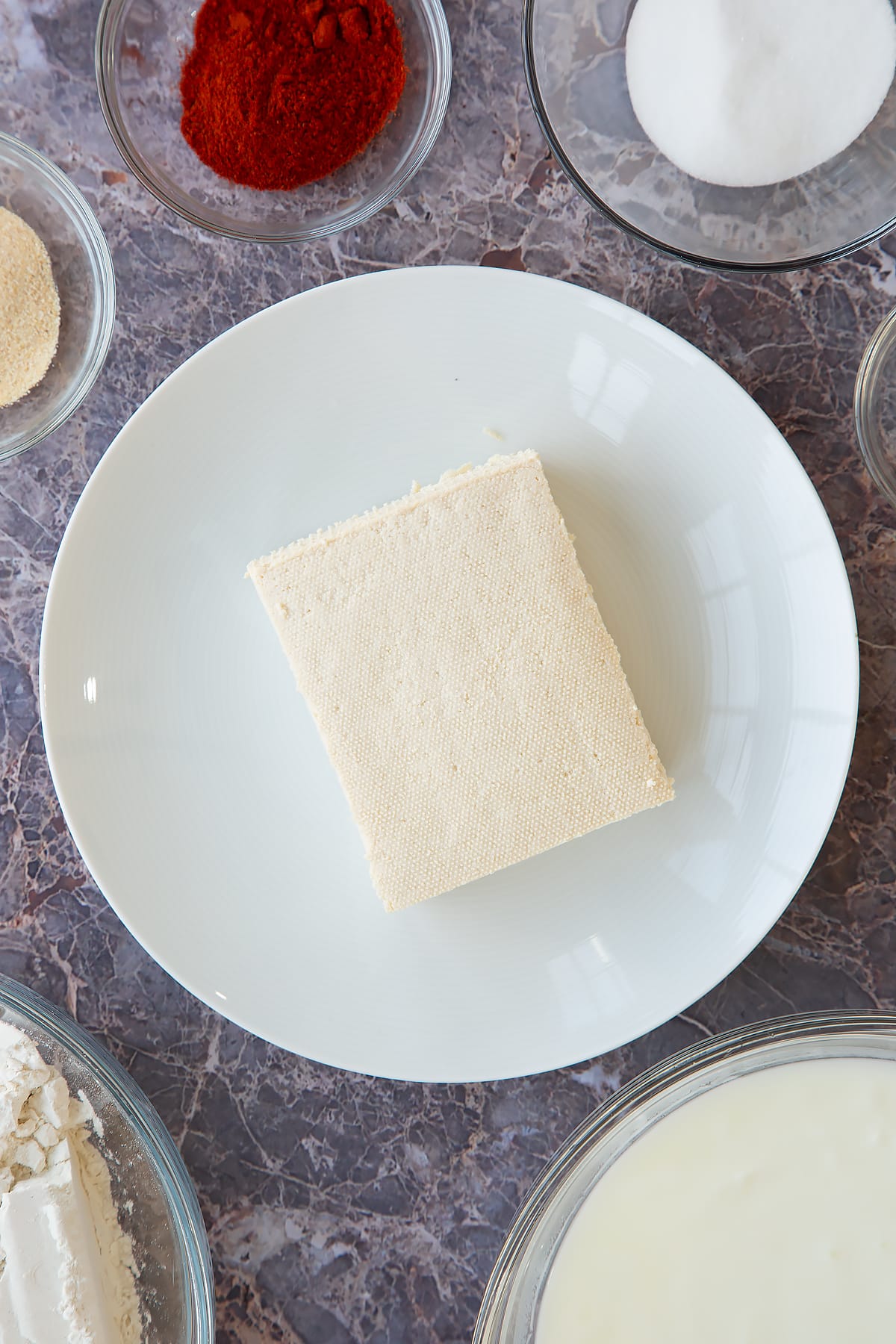 a block of tofu on a white plate surrounded by spices and herbs.