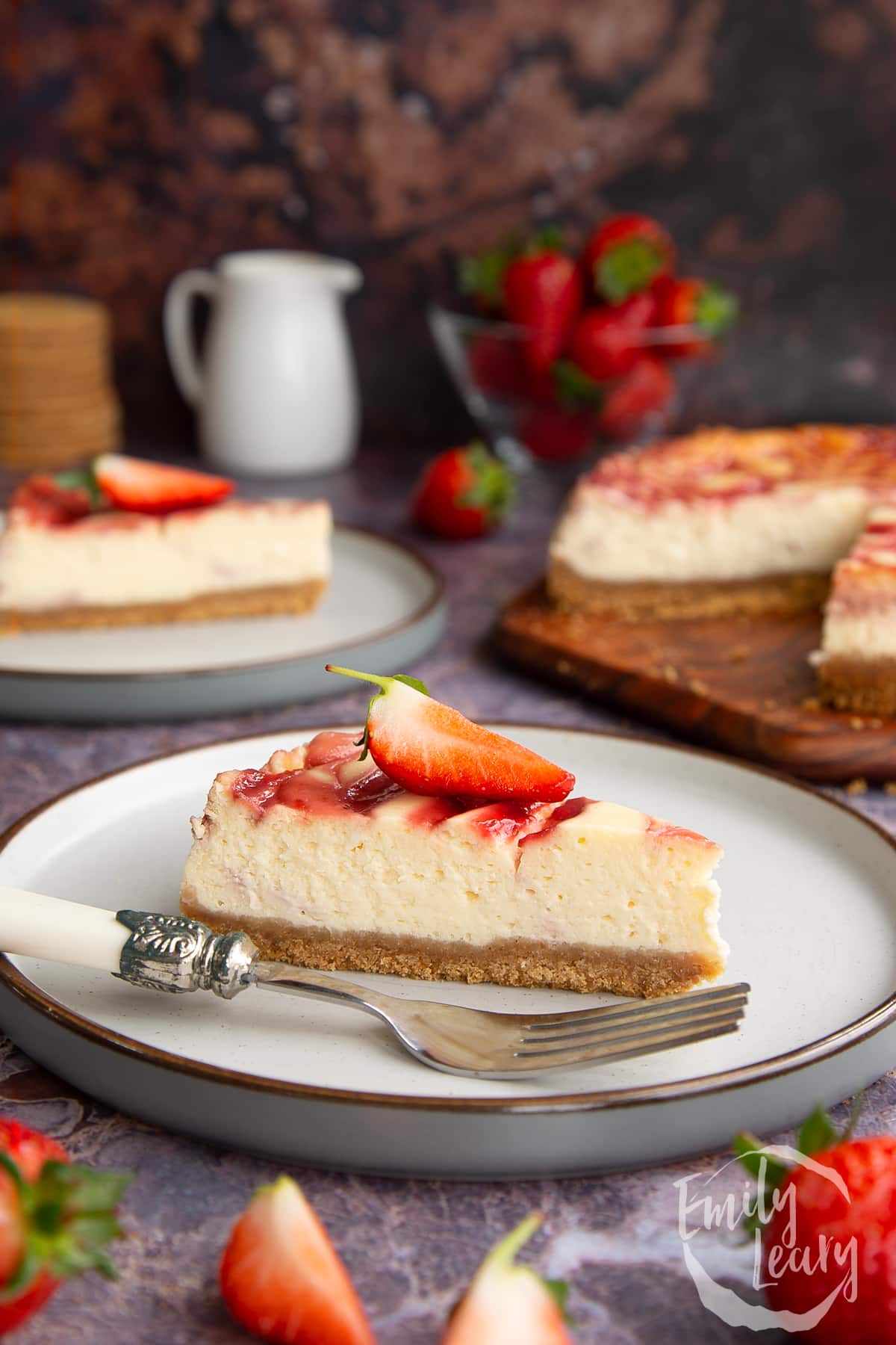 a slice of Strawberry swirl cheesecake on a white plate with a fork on front topped with a cut strawberry.