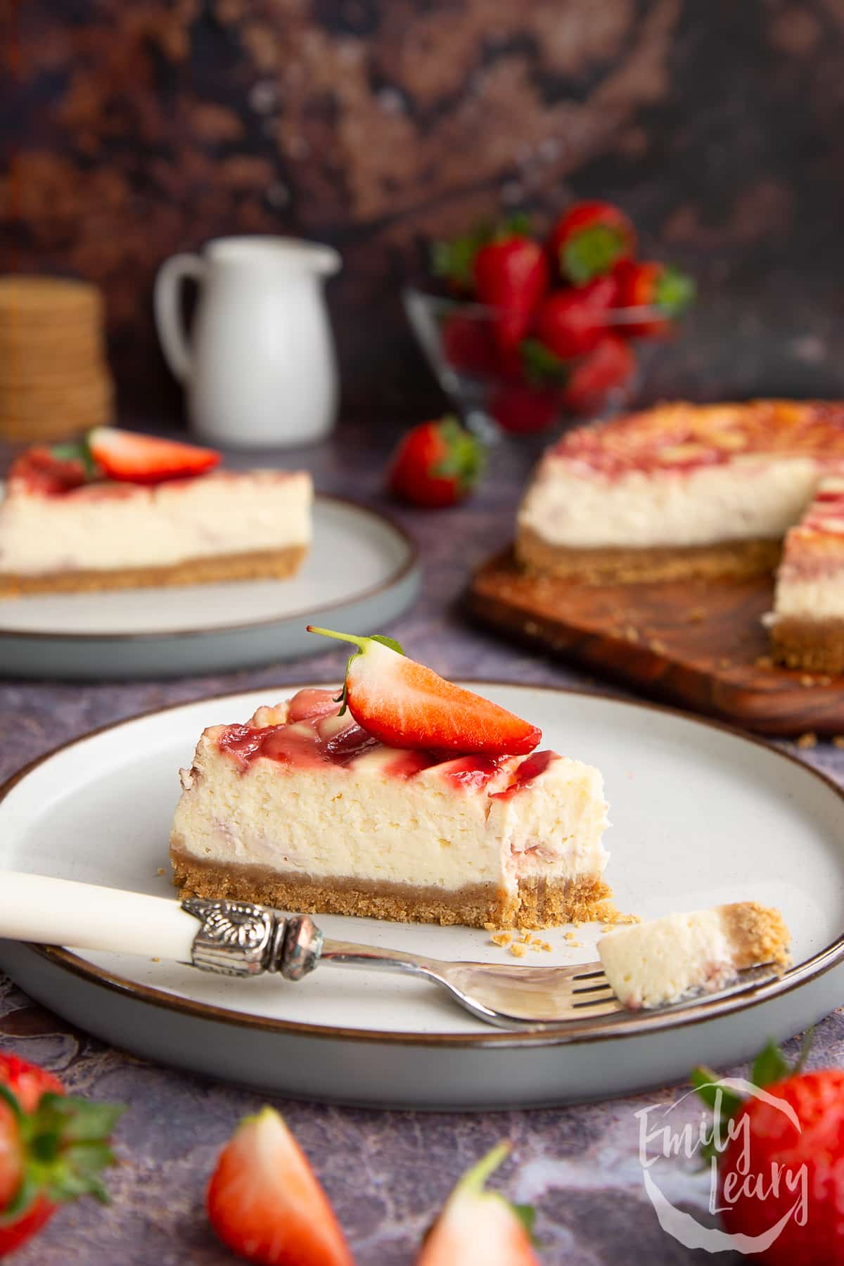 a slice of Strawberry swirl cheesecake on a white plate with a fork on front topped with a cut strawberry and a bite missing from the front.