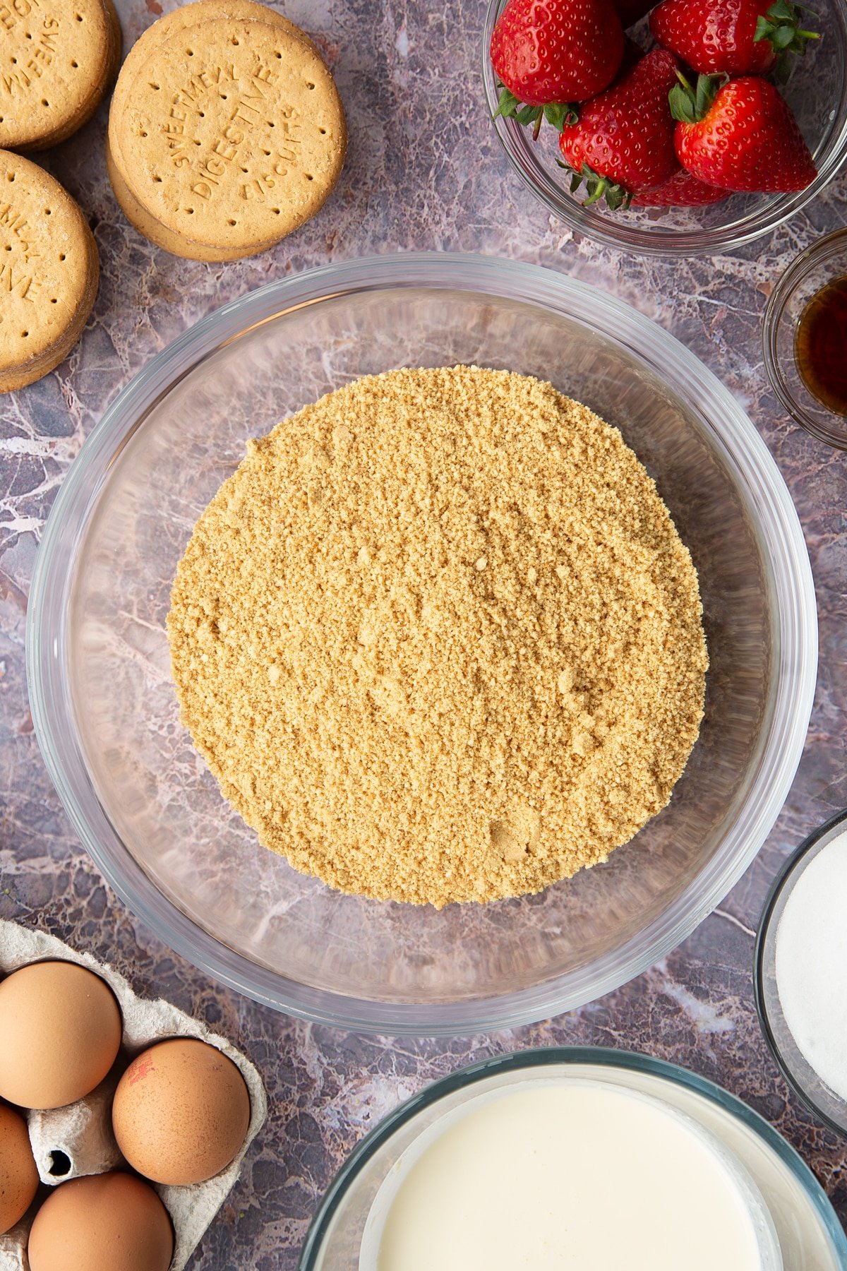 crushed digestive biscuit crumbs in a large clear bowl surrounded by digestive biscuits and strawberries.